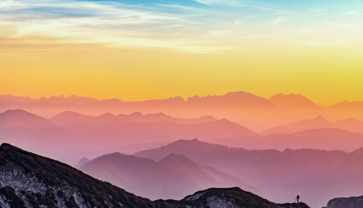 Photo of colourful sunset over mountains. Very small person hiking in the foreground.