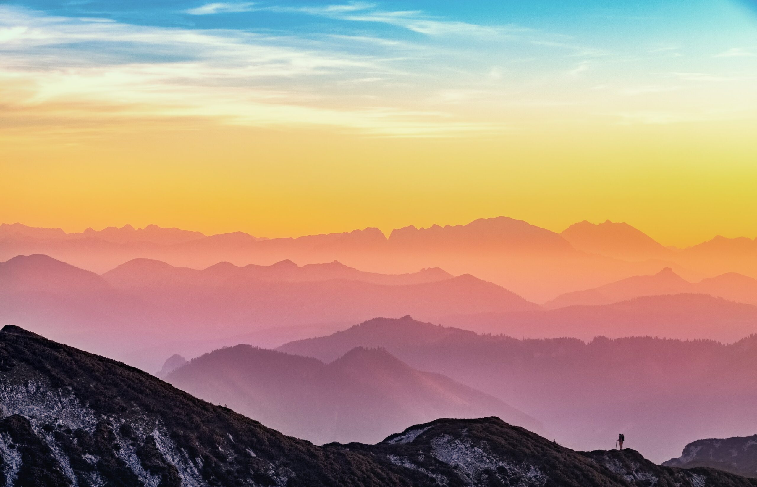 Photo of colourful sunset over mountains. Very small person hiking in the foreground.
