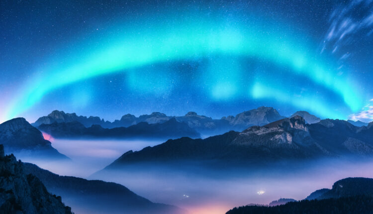 Aurora borealis above mountains in fog at night. Northern lights. Sky with stars with polar lights and high rocks. Beautiful landscape with aurora, city lights in low clouds, mountain ridge
