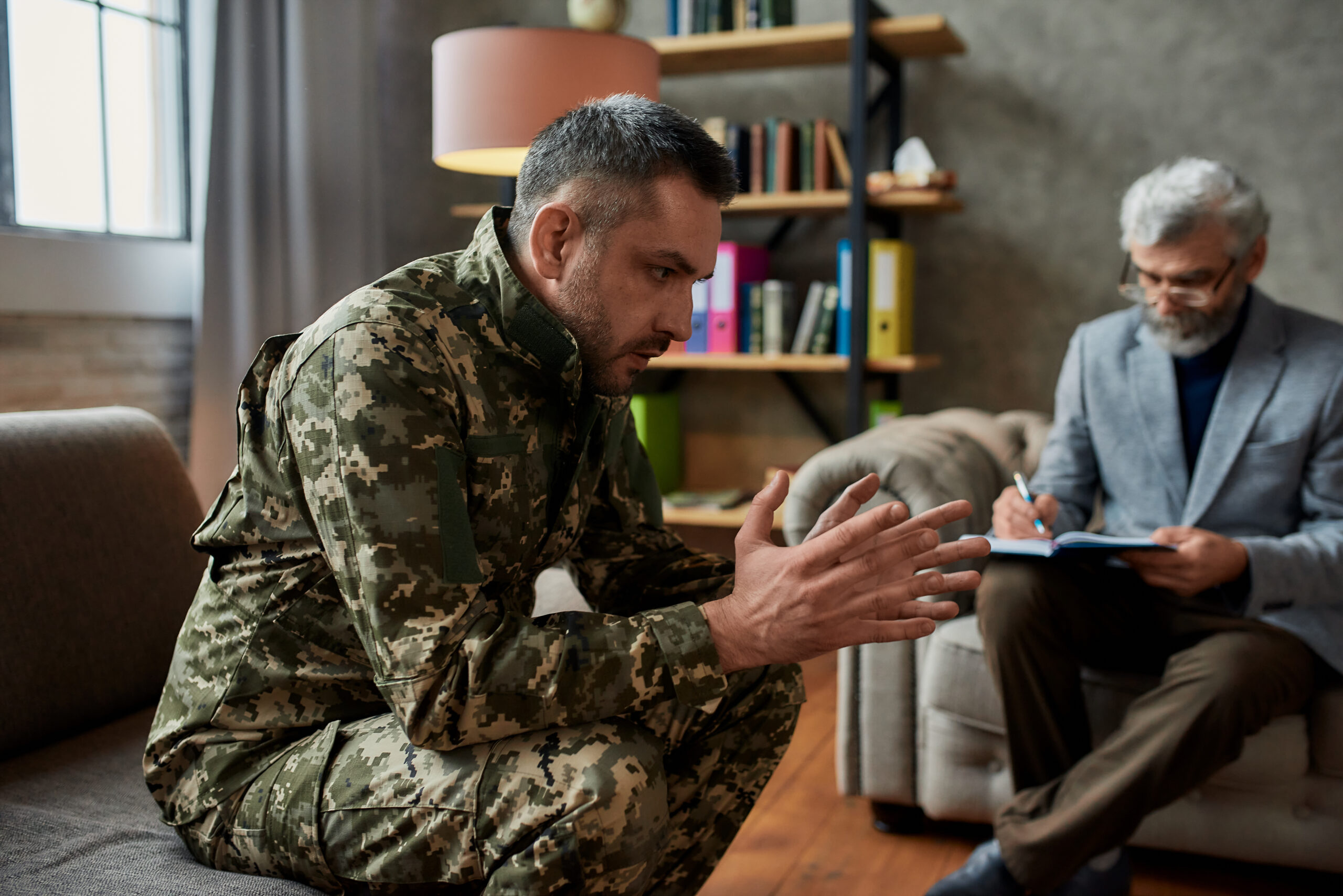 Thoughtful middle aged military man communicating with psychologist, sitting on the couch during therapy session. Soldier suffering from depression. PTSD concept. Horizontal shot. Focus on military