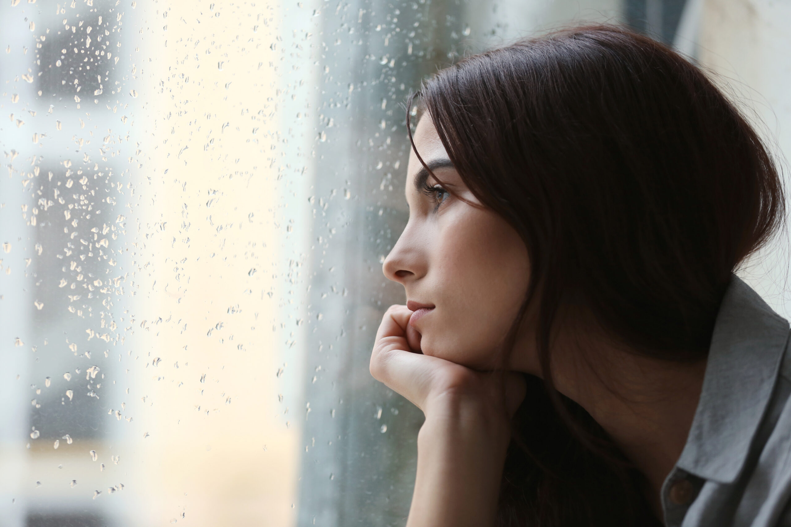 Depressed woman with dark brown hair looking out window. It's raining outside.